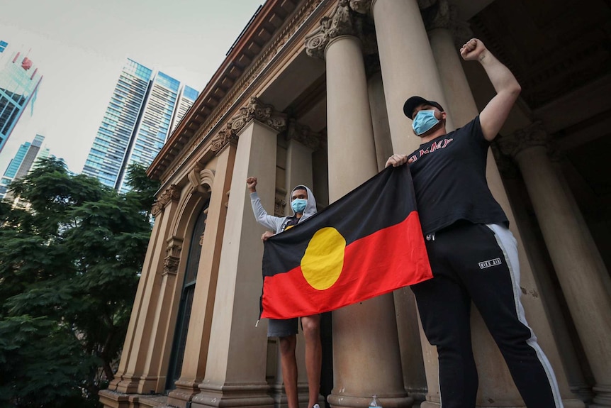 Two men carrying an Aboriginal flag raise their clenched fists.