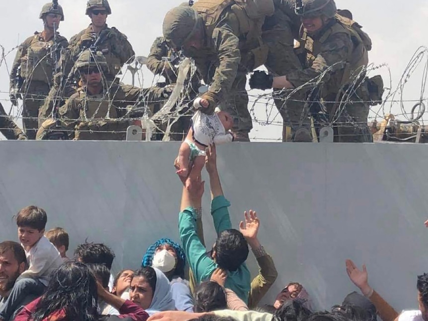 A soldier on a wall grabs the arm of a baby held up by a civilian