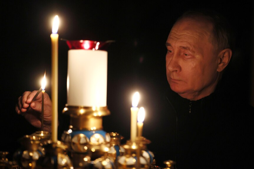 Putin standing in the dark wearing black lighting a candle on a gold church candle holder