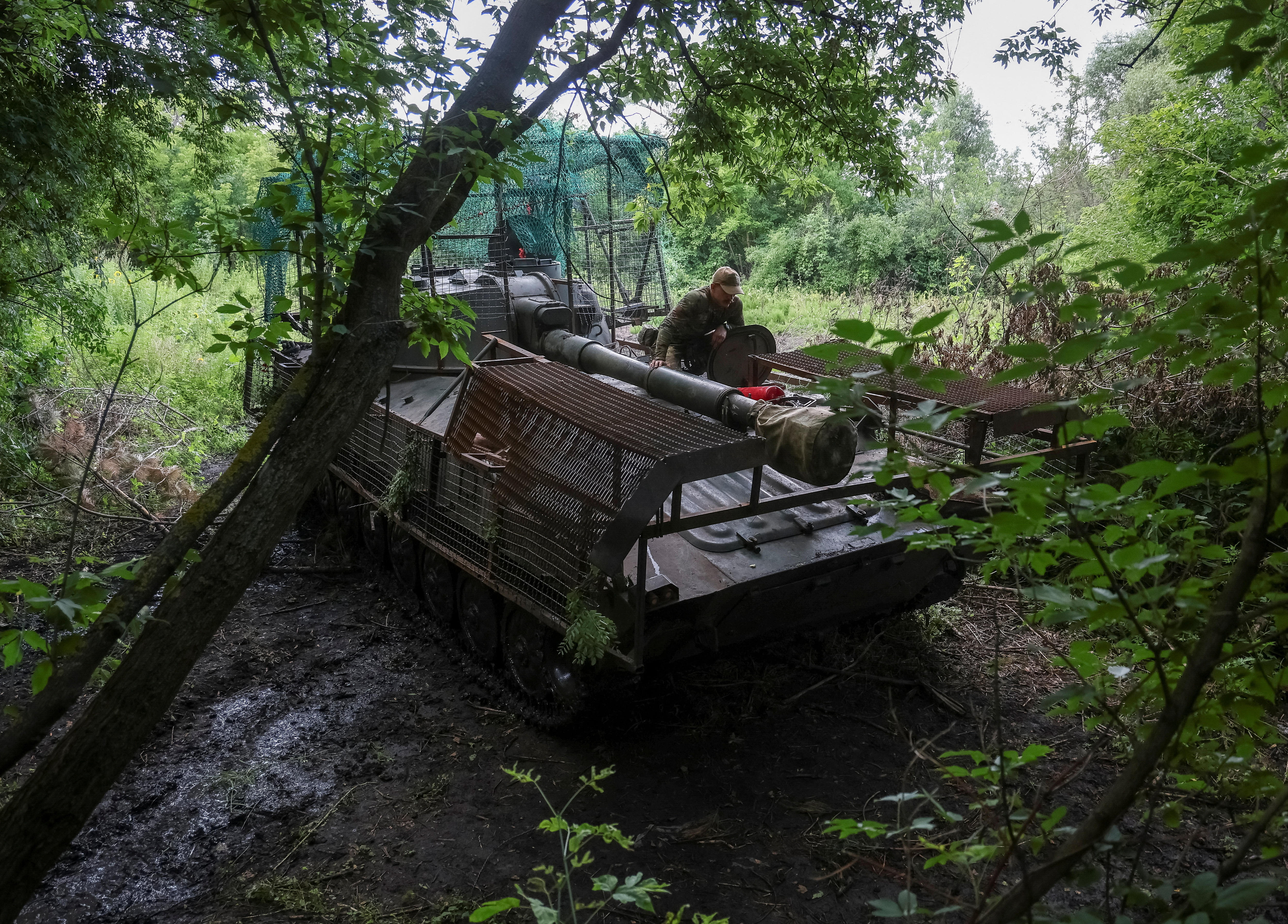 A Ukrainian serviceman sits on top of a self-propelled howitzer beneath a canopy of trees.