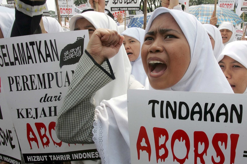 A woman wearing a white Islamic veil yelling while holding a sign, surrounded by other Muslim women.