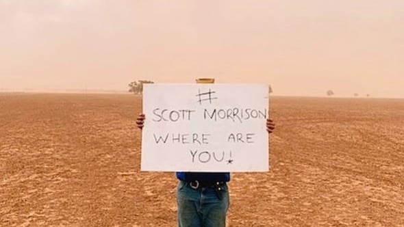 A man holds a sign reading "scott morrison where are you" as he stands in the middle of a bare and dusty field