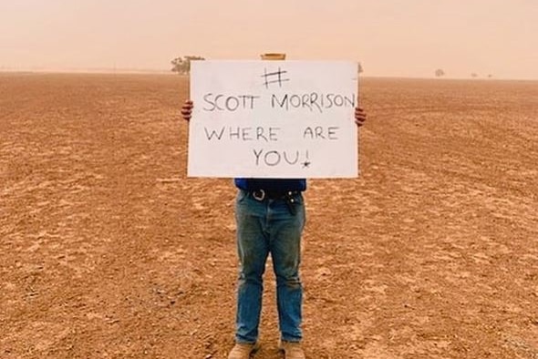 A man holds a sign reading "scott morrison where are you" as he stands in the middle of a bare and dusty field