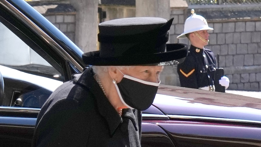 A close-up of the Queen shows her n black mourning attire, with a black hat and face mask. 