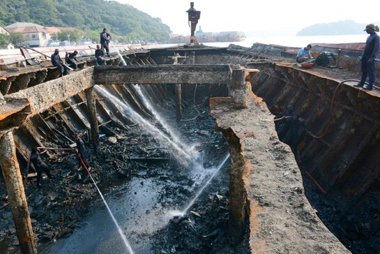 A crew of workers stands on the rim of a rusted shipwreck, some of them squirting hoses inside