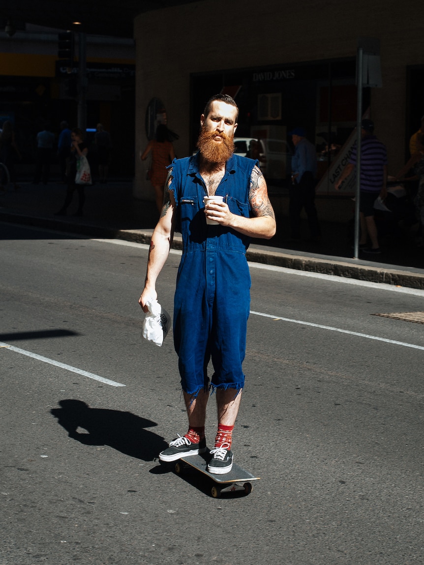 A bearded man in cut-off overalls rides a skateboard in Sydney.