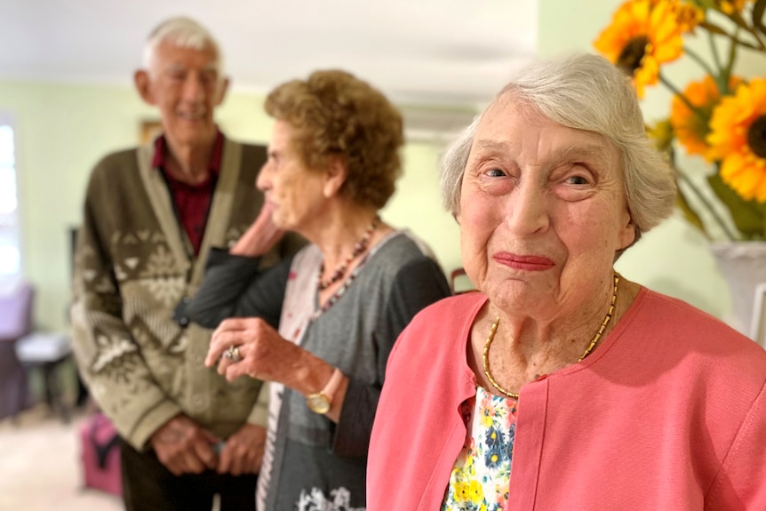 Woman in her 80s grins at the camera with an older couple in the background