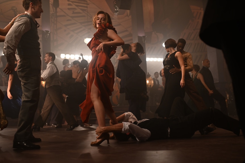 A young woman in a red dress dances in a retro club setting, hiking her skirt up, with a man on the floor grasping her ankles