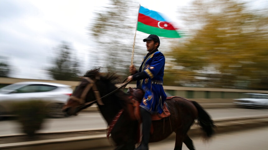 You view a man on a horse waving a Azerbaijani flag who is pictured mid-motion while riding down a main road.