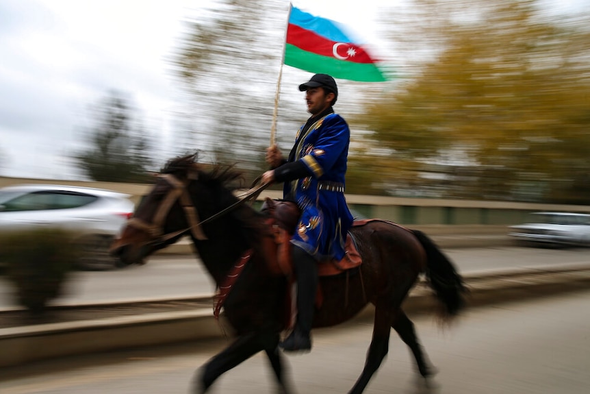 You view a man on a horse waving a Azerbaijani flag who is pictured mid-motion while riding down a main road.