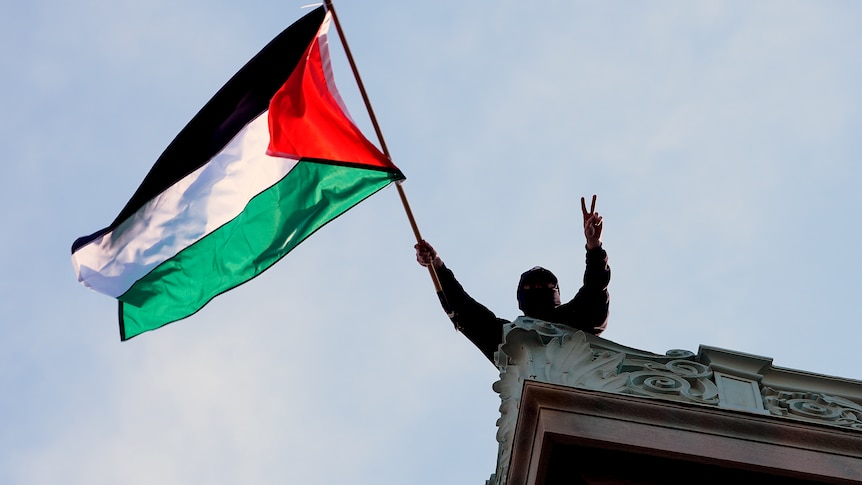 A protester on the roof of a building holds up the peace/victory sign and waves a Palestinian flag
