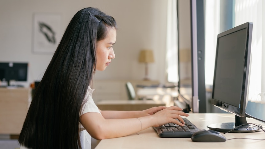 A young women with long black hair types on a computer in a well-lit office.