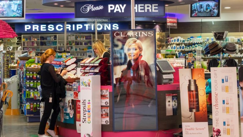 A woman stands at the counter of a pharmacy surrounded by products and advertising.