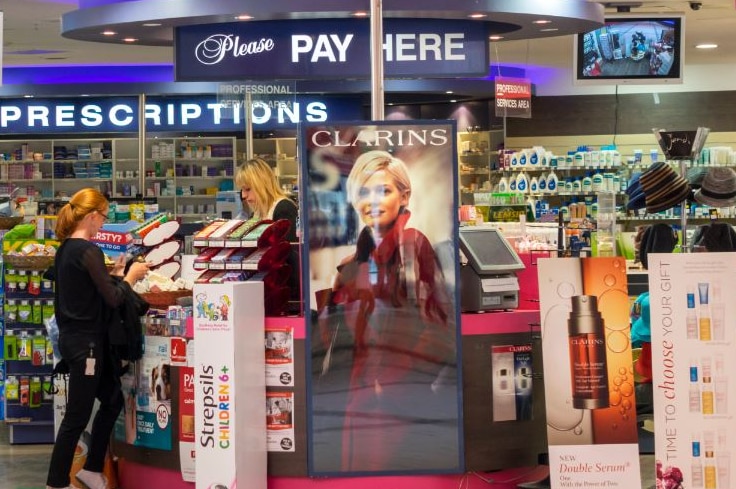 A woman stands at the counter of a pharmacy surrounded by products and advertising.