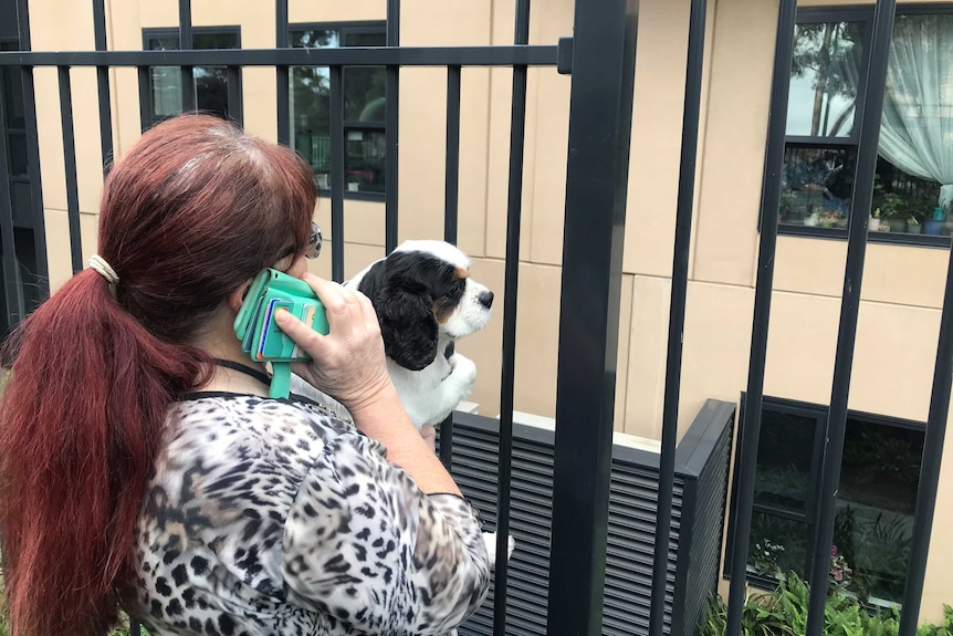 A woman stands at a fence and speaks on the phone while holding a dog