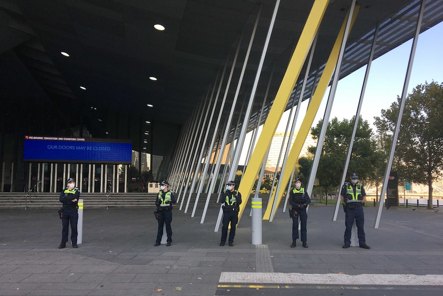 Five police officers wearing face masks stand about 2 metres apart in a line outside a convention centre.