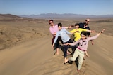 Five people stand on a sand dune with their hands raised before a dry desert basin, with mountains in the distance 