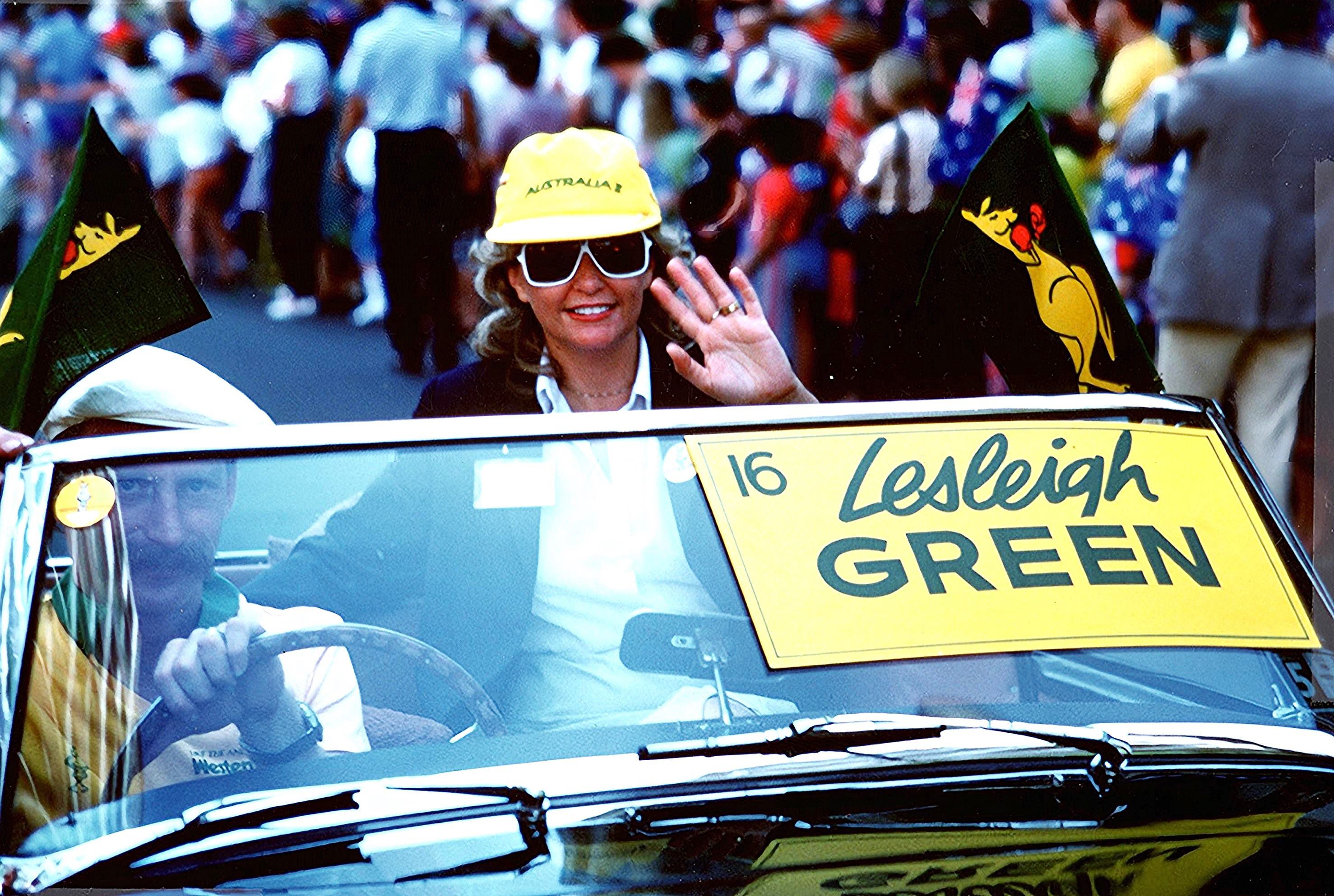 A smiling woman waves from her seat in a convertible car bearing a sign saying "Lesleigh Green" on its windcreen.