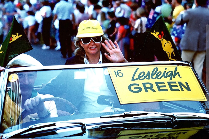 A smiling woman waves from her seat in a convertible car bearing a sign saying "Lesleigh Green" on its windcreen.