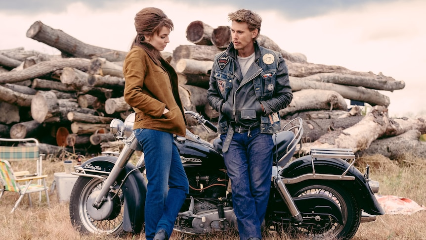 A young man and woman talk in front of a motorbike and a pile of logs