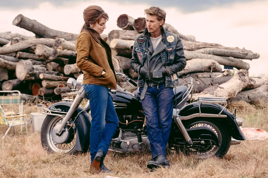A young man and woman talk in front of a motorbike and a pile of logs