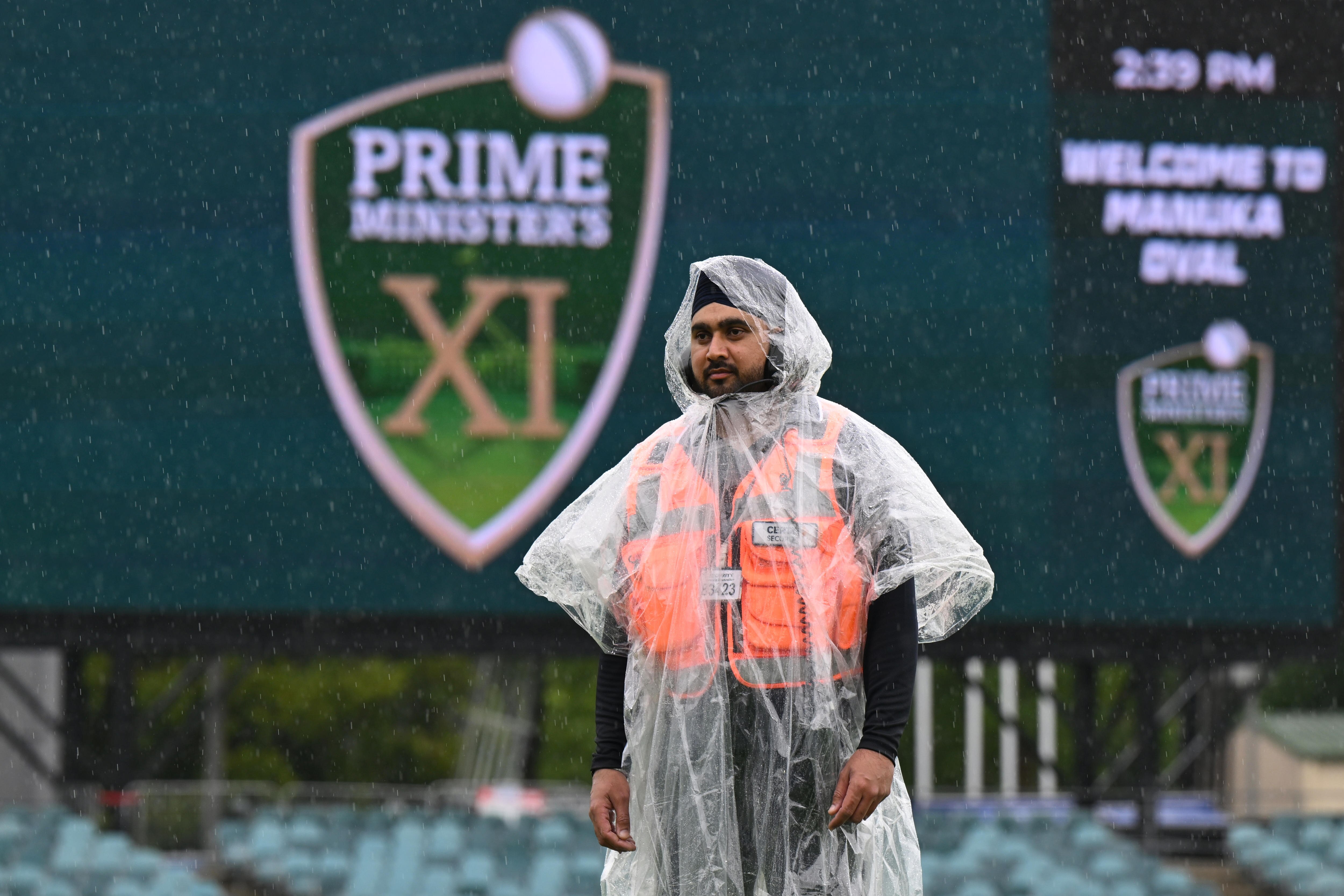 A security guard wears a rain poncho at Manuka Oval with Prime Minister's XI displayed on the screen behind him.