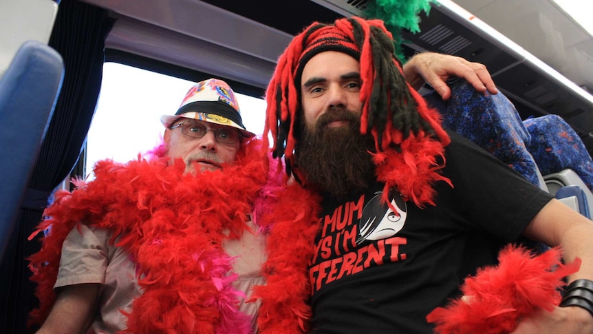A man in a colourful hat and red feather boas with his arm around another man in a red and black wig.