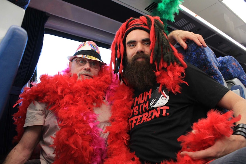 A man in a colourful hat and red feather boas with his arm around another man in a red and black wig.