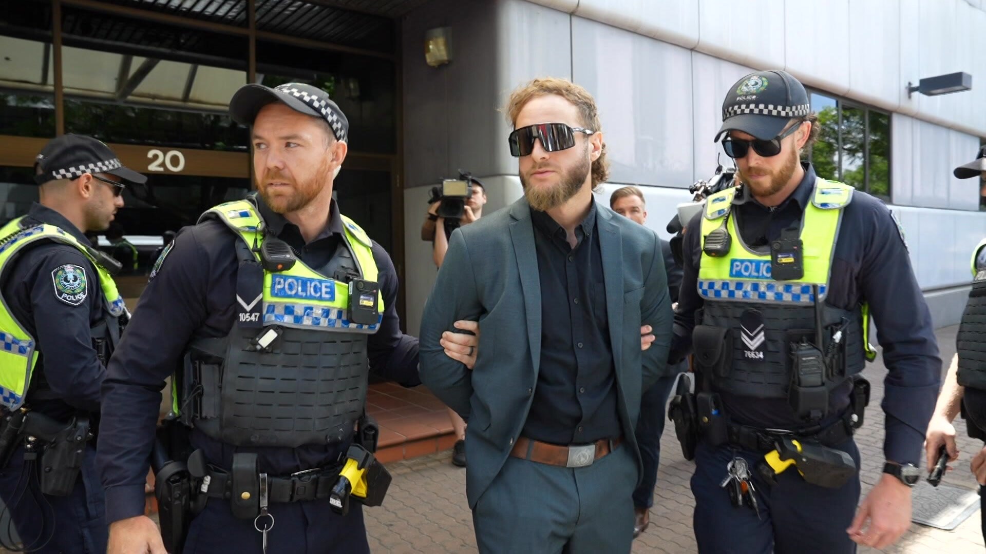 Joel Davis - man with mullet and beard wears green suit jacket and large dark sunglasses taken away by police