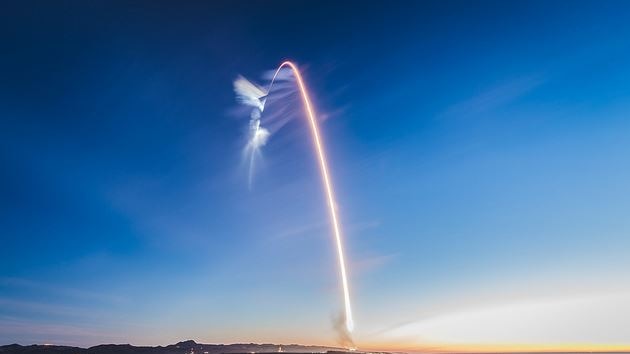 A rocket launch viewed from the distance, with the rocket's trail forming an arc in the sky.