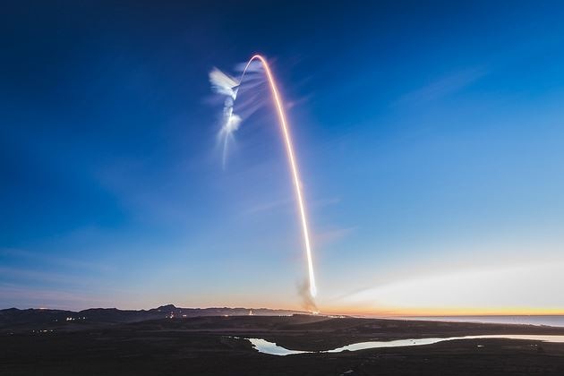 A rocket launch viewed from the distance, with the rocket's trail forming an arc in the sky.