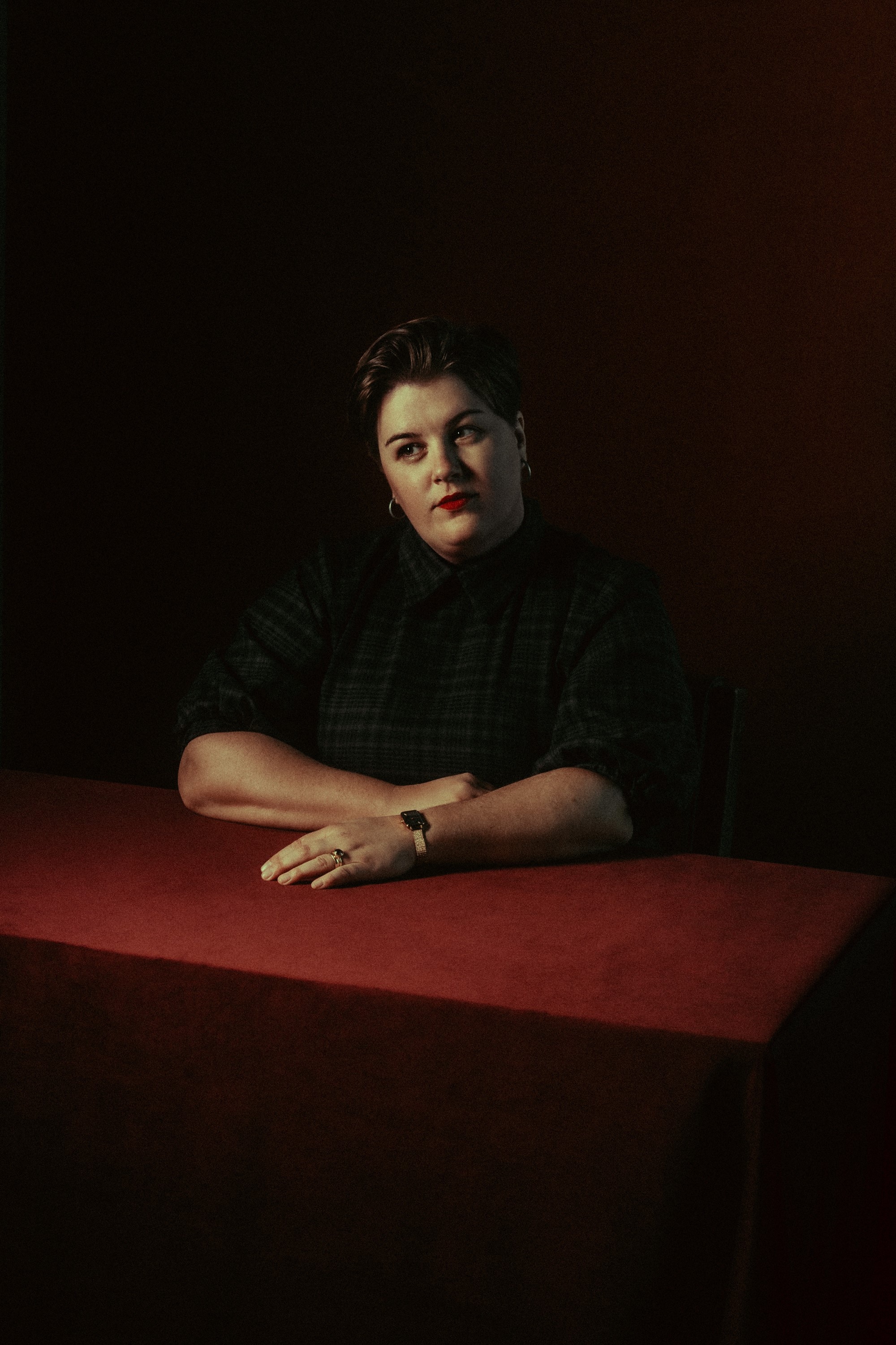 A person with a black buttoned up shirt, a red lip, and their arms folded in front of them on a table