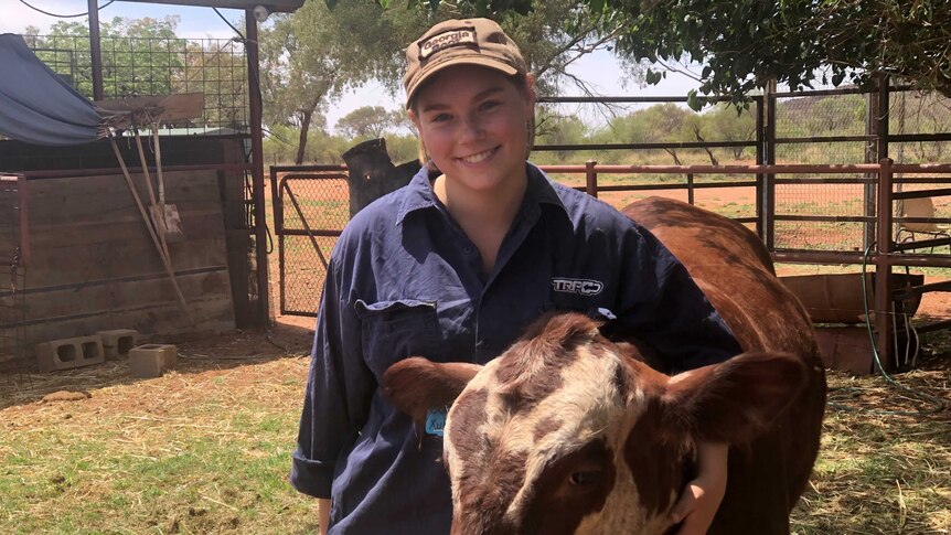 A young woman stands with her arm around a cow in a cattle yard, smiling.