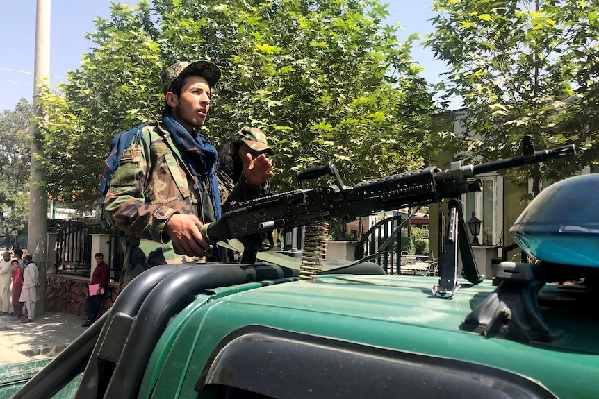 A middle-eastern soldier in army fatigues holds a gun as he stands above a green vehicle on city street.