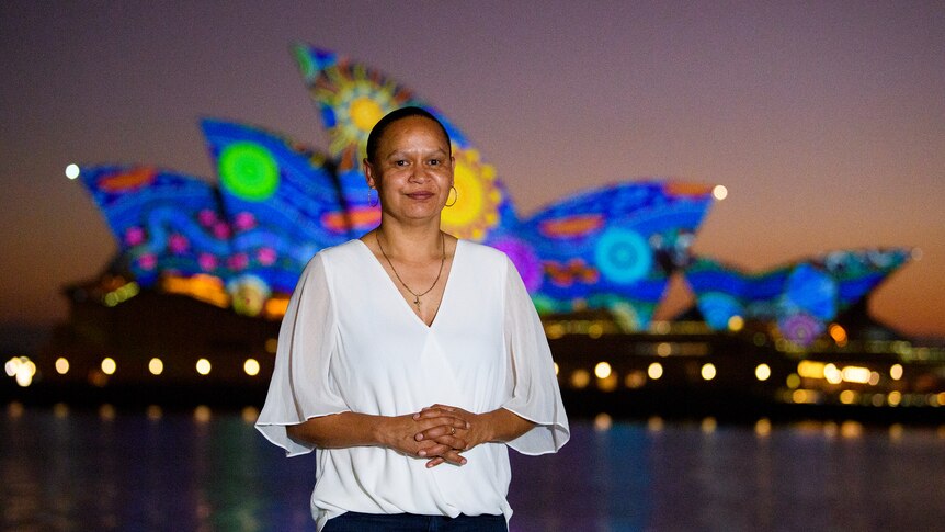 a woman stands in front of the illuminated Sydney Opera House sails at dawn