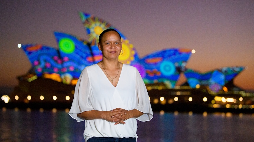 a woman stands in front of the illuminated Sydney Opera House sails at dawn