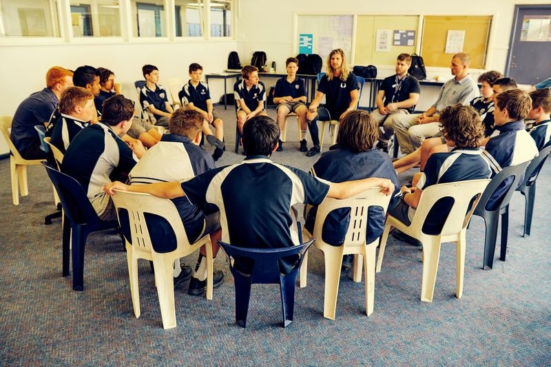 A group of young male students sit iwth two older men in a circle on plastic chairs inside a classroom.