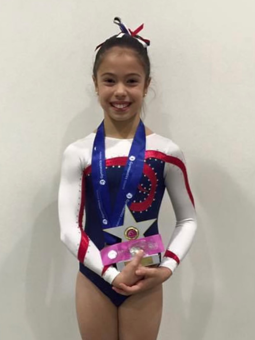 Scarlett Magnanini stands in front of a wall in her gymnastics leotard holding medals.