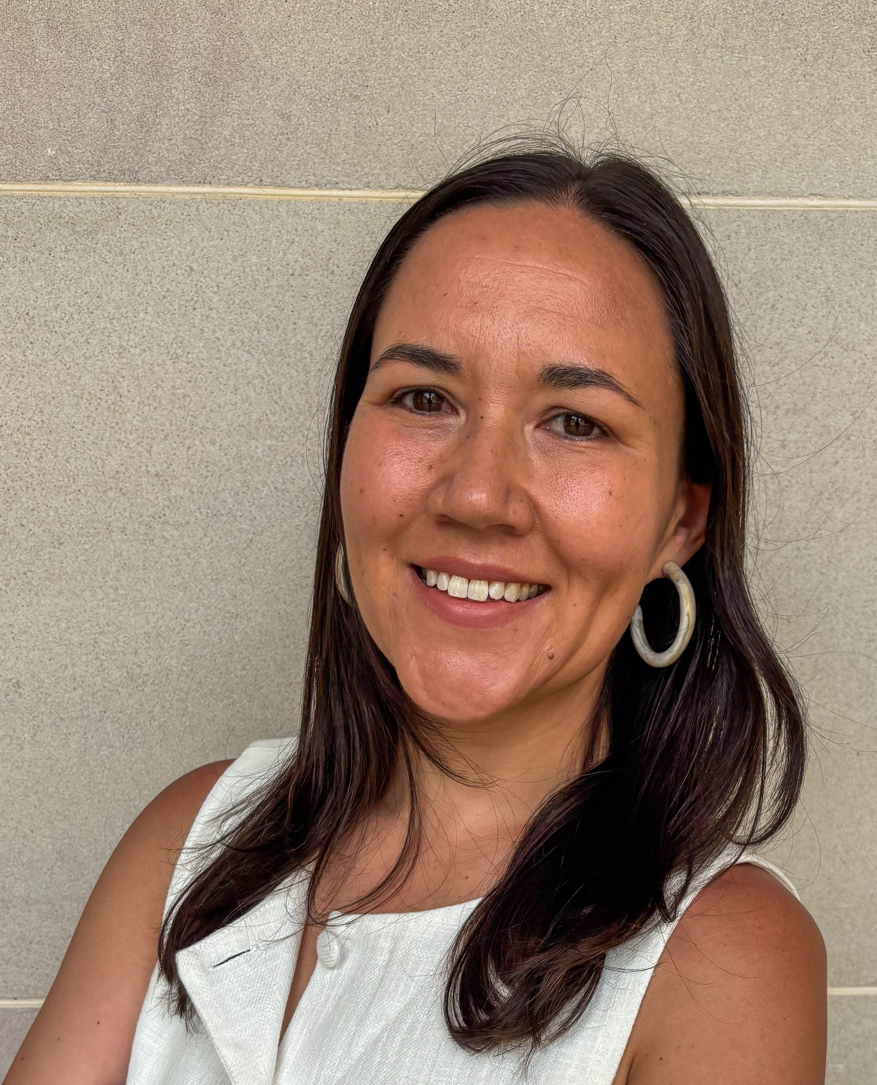 A portrait of Tegan Cohen. She is smiling at the camera, has long dark hair and is wearing hoop earings.