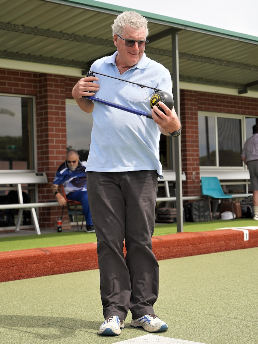 A man on a bowling green cradling a ball with the aid of an arm-extending device.