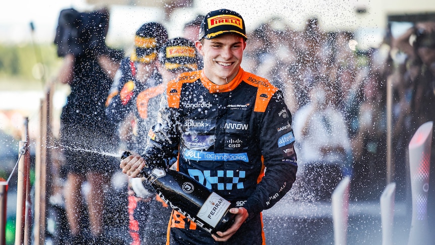 An F1 driver wearing orange and black, sprays wine on the podium and smiles