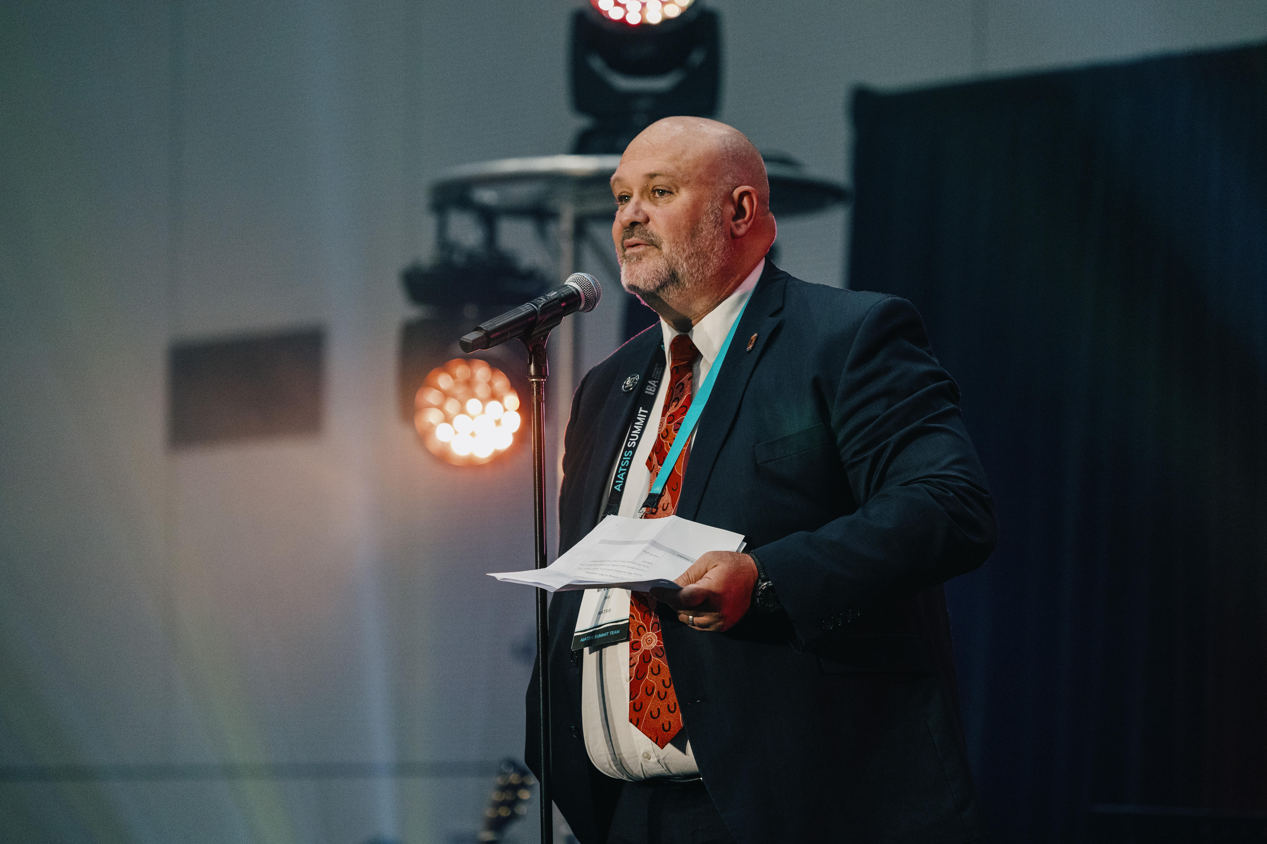 AIATSIS CEO Leonard Hill stands on stage in a blue suit talking into a microphone