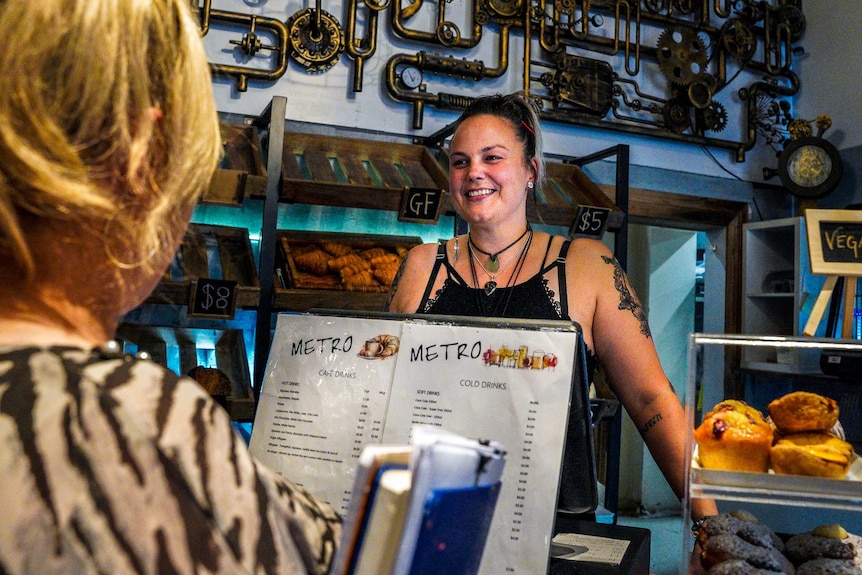 A young woman smiles in a cafe interior as another woman looks on.