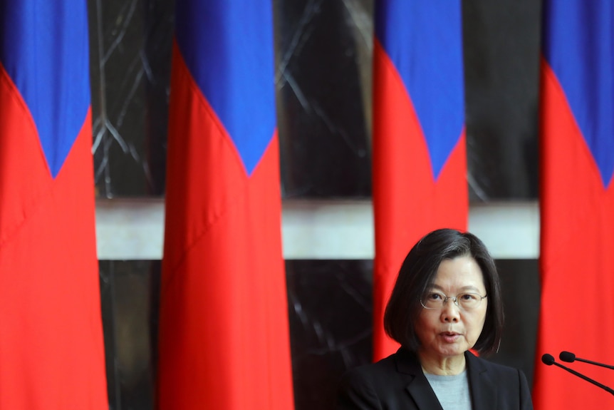 Tsai Ing-Wen, in a black blazer, stands with microphones in front of red and blue Taiwanese flags