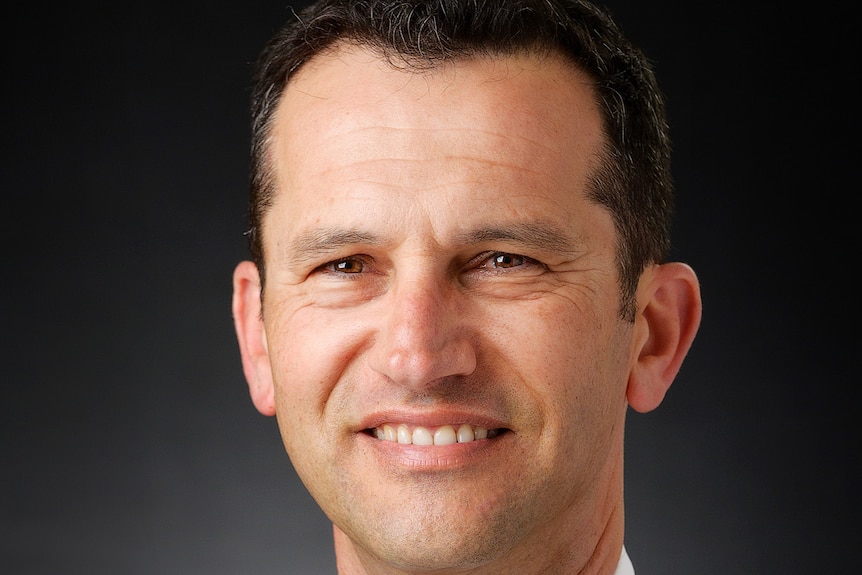 A headshot of a man with dark hair wearing a blue suit and yellow and navy striped tie