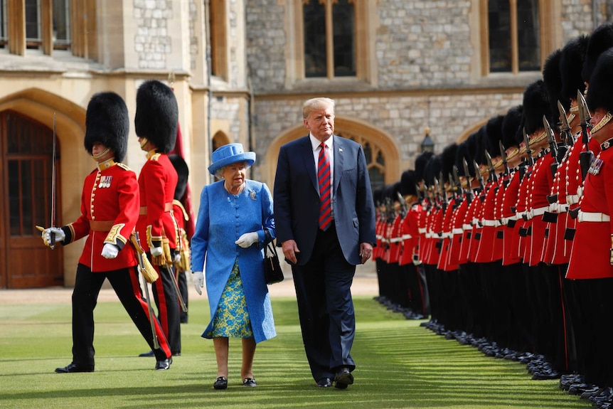 Queen dressed in blue walks next to Trump on green lawn past a line of guards in red jackets holding bayonets.