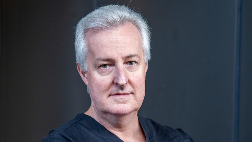 grey haired man with arms crossed wearing blue medical scrubs