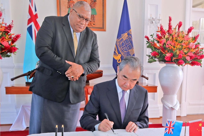 Fiji's President Ratu Wiliame Katonivere stands behind China's Wang Yi as sits and signs a document.