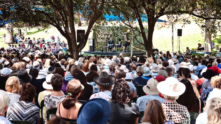 A large crowd of people gather under trees to watch some people sitting on a stage. 