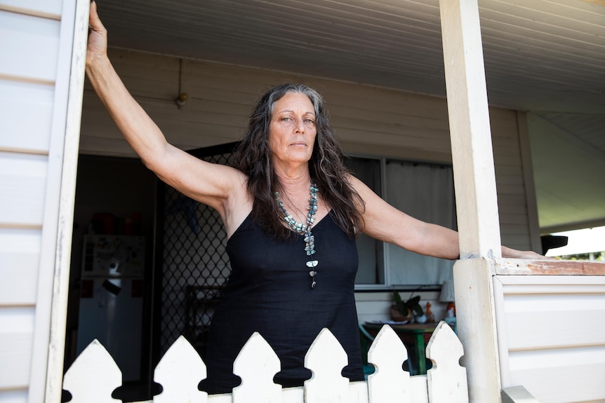 A woman with wavy brown hair, wearing a black dress, stands on the patio of a white weatherboard house.